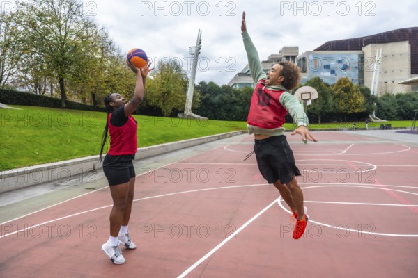 Young diverse friends are actively playing basketball on an outdoor court, a man jumping high attempting to block a woman shooting the ball, emphasizing teamwork, competition, and dynamic sport