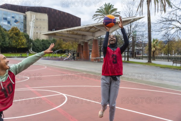 Woman in hijab smiling and shooting a basketball during a friendly game on an outdoor urban court, with a man defending her and buildings in the background