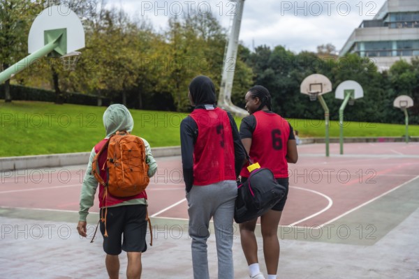 Young diverse friends wearing sports bibs and carrying bags, walking united towards basketball hoops on an outdoor court, ready for a game or sports practice