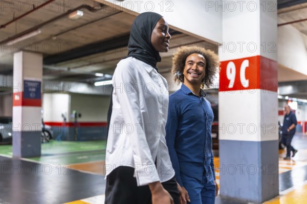 Diverse young couple walking happily and smiling together in a modern underground parking garage after parking their vehicle, representing urban transportation and connection