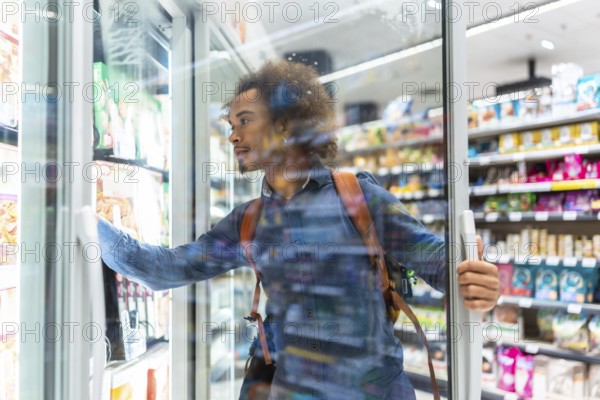 Young black man browsing frozen food in a supermarket aisle, opening a freezer door and selecting packaged items for meal prep and grocery shopping