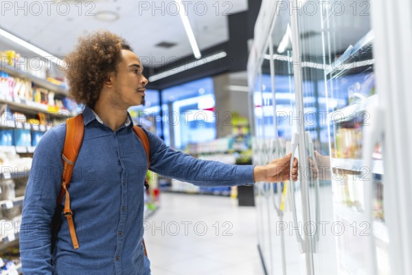 Young man with backpack opens supermarket freezer and selects products from a refrigerated aisle, casually choosing frozen and fresh items during everyday shopping trip