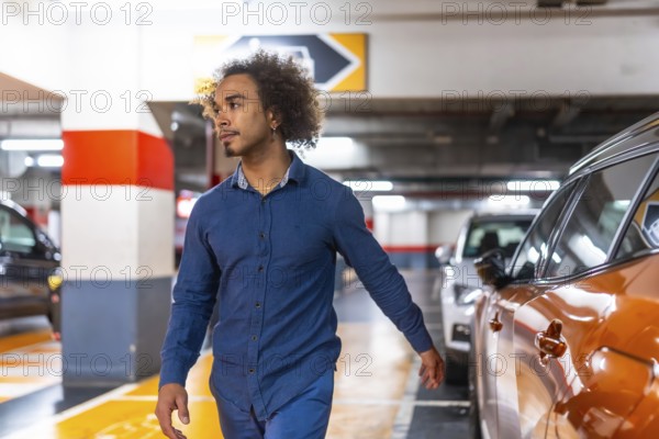 Young man with afro hair walks through a brightly lit underground parking garage, glancing right as he searches for his car among rows of vehicles and concrete columns
