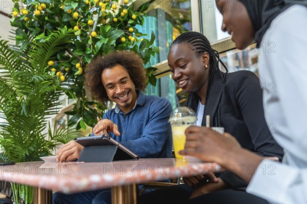 Diverse group of young adult friends enjoying a social gathering in a modern cafe, laughing and collaborating while interacting with a digital tablet on the table