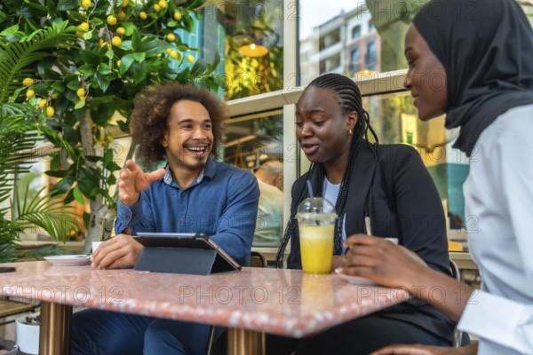Diverse young professionals and friends gathered in a relaxed cafe, laughing, sharing ideas and collaborating over drinks while using a tablet for brainstorming and teamwork