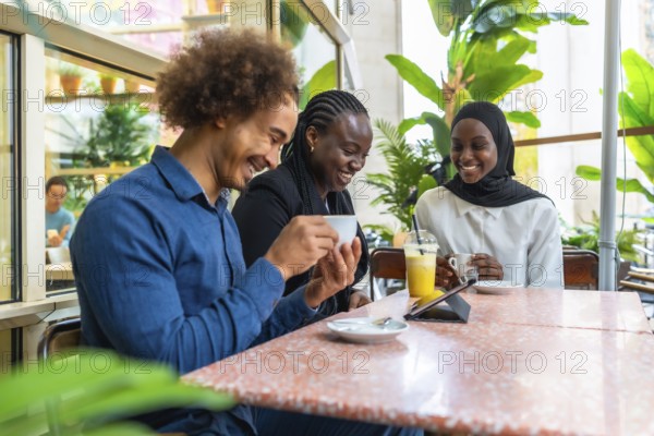Diverse group of friends and coworkers gathered in a modern cafe, smiling and laughing over coffee and juice while sharing a digital tablet and enjoying a relaxed meetup