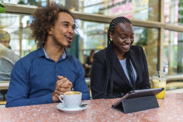 Two diverse business professionals are meeting in a relaxed cafe environment, discussing ideas and collaborating while using a tablet and enjoying beverages