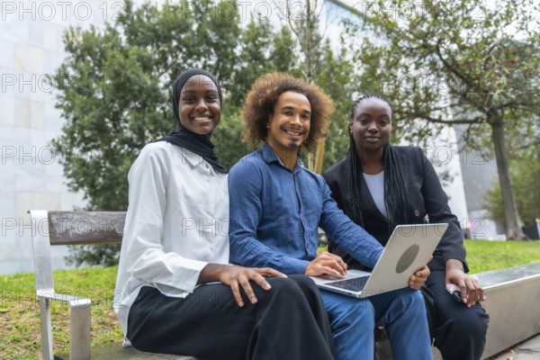 Three diverse students are smiling directly at the camera while sitting outside on a bench. Working together on a laptop. Symbolizing teamwork. Education. And modern learning in a relaxed environment