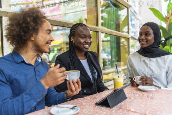 Diverse group of young adult friends smiling and engaging in conversation while sitting at a table in a modern cafe, drinking coffee and juice alongside a tablet