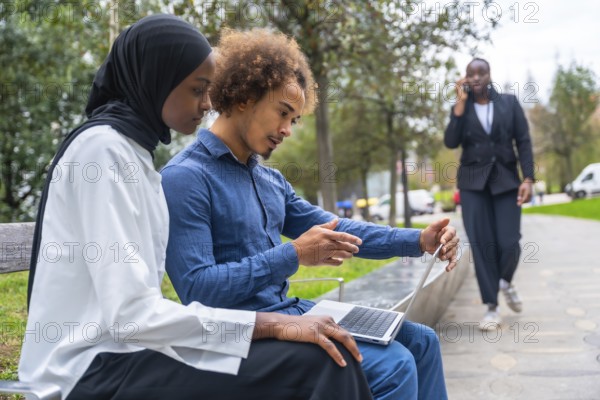 Young diverse colleagues collaborating on a portable computer in an urban park, with a woman in the background walking and talking on her mobile phone, highlighting remote work and tech integration