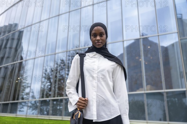 Confident young black woman wearing a hijab and carrying a shoulder bag, standing in front of a modern glass building, representing education, university life, and ambition