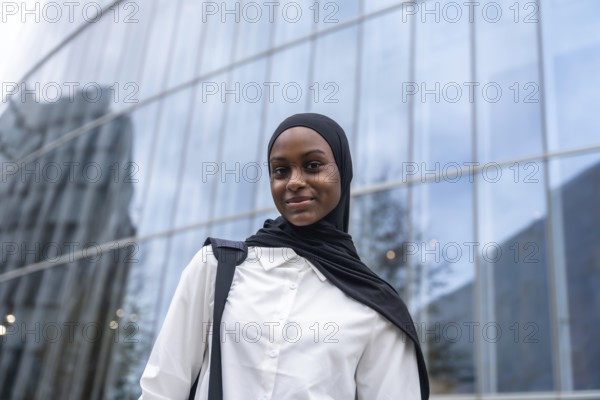African american woman student wearing a black hijab and white shirt, carrying a backpack, standing confidently in front of a modern glass building in an urban environment