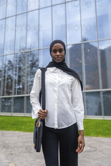 Young black muslim woman wearing a hijab and white shirt, holding a bag and standing confidently outside a contemporary glass building, representing education and successful futures