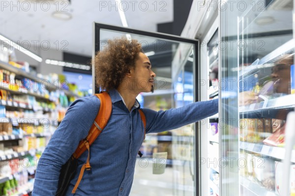 Young man shopping in a grocery store, reaching for frozen items in a refrigerated display, reflecting modern consumer behavior, food selection, and convenience in daily life