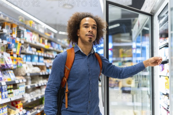 Young man with backpack selecting chilled items from a refrigerated display in a bright modern supermarket aisle, thoughtful consumer choosing fresh grab and go options