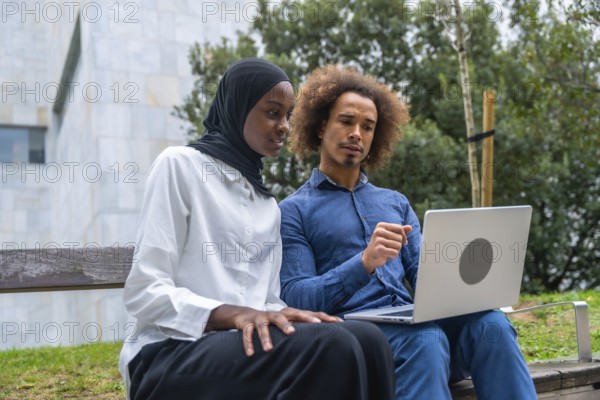 Diverse young man and woman collaborating on a laptop while seated on a park bench, focused on work or studying together in a sunny urban green space, casual and professional