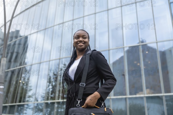 Young black businesswoman smiling and holding a laptop bag, walking with confidence outside a modern glass office building, symbolizing success and career progression in the corporate world
