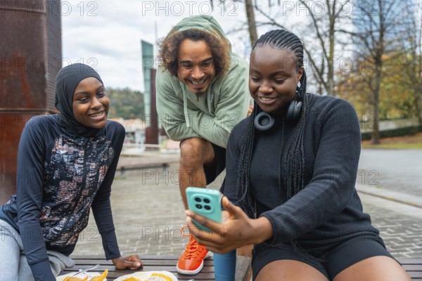 Three diverse young adults, a woman in a hijab, a man with curly hair, and a woman with braids, smiling and sharing content on a smartphone outdoors in a park