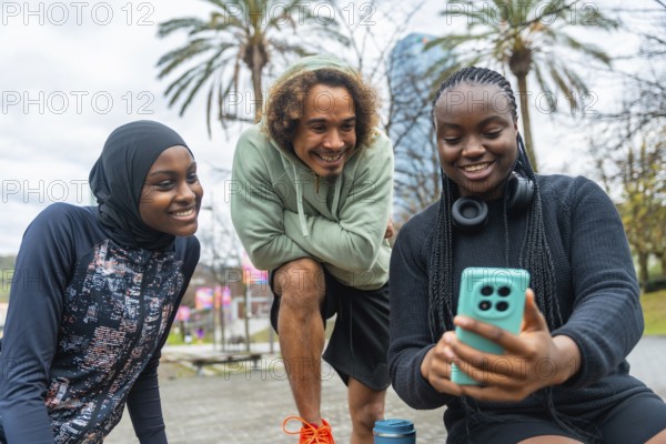 Group of diverse young friends, including a woman wearing a hijab, bonding and sharing a joyful moment while looking at a smartphone screen in a park setting