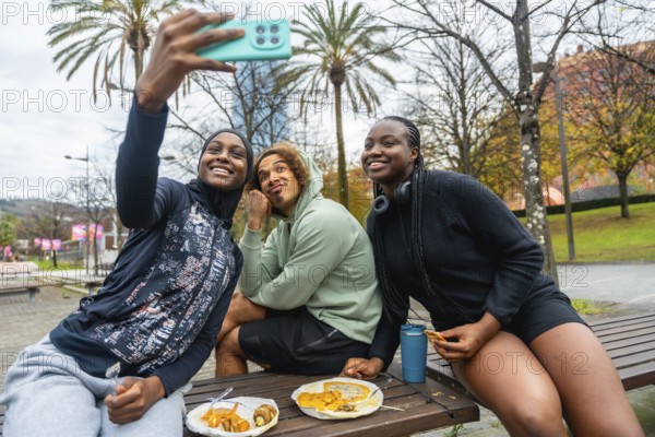 Diverse group of young adult friends smiling while taking a cheerful selfie and eating take out food outdoors in a public park, sharing a moment of leisure