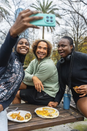 Diverse group of young adult friends smiling and laughing, holding a phone to take a selfie while enjoying a casual picnic with food and drinks on a wooden bench in an urban park