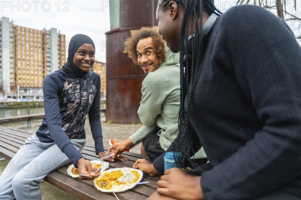 Diverse group of young adults sharing a casual outdoor meal on a city park bench, smiling, talking and relaxing together during an urban lunch or picnic break