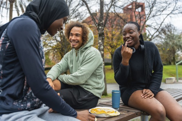 Group of diverse african friends happily laughing and enjoying a meal together on a park bench, sharing moments of leisure and conversation in an urban park setting