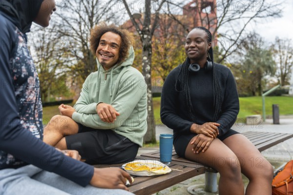 Group of diverse young adults sitting on a park bench, sharing a meal and enjoying casual conversation outdoors, fostering friendship, connection, and wellbeing