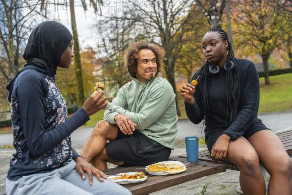 Group of diverse friends sitting on a park bench, sharing a casual picnic meal outdoors while enjoying conversation and relaxing after physical activity, promoting friendship and inclusivity
