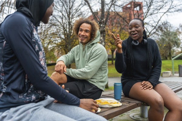 Group of diverse young adult friends sitting on a park bench, engaging in cheerful conversation and sharing a meal while enjoying leisure time outdoors