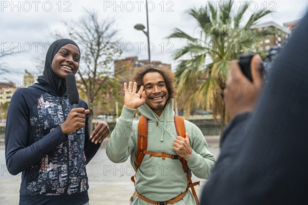 Smiling female journalist wearing a hijab holding a microphone, interviewing a happy man waving on the street while filming for a news report, capturing diverse content