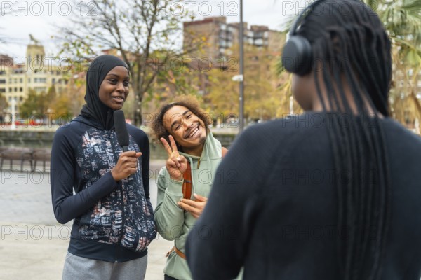Young black woman wearing a hijab interviewing a smiling friend making a peace sign, while another friend with headphones listens to the discussion outside in an urban park