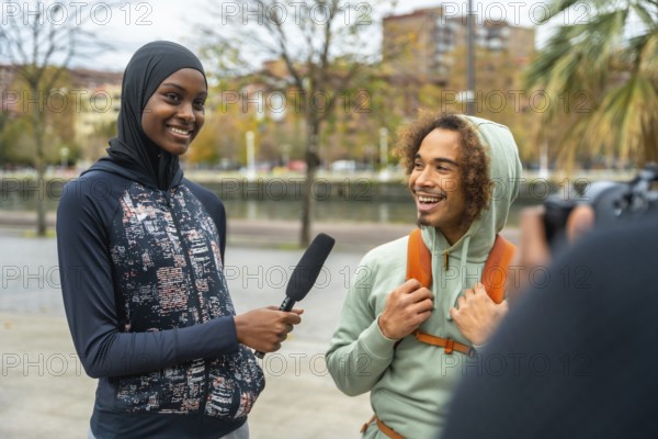 Female journalist wearing a hijab holding a microphone, interviewing a cheerful young man with curly hair in a vibrant city environment while filming the conversation