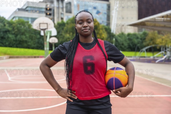 Confident young black woman athlete with braids wearing a red jersey, holding a basketball, and standing on an outdoor court with hands on hips, looking directly at the viewer