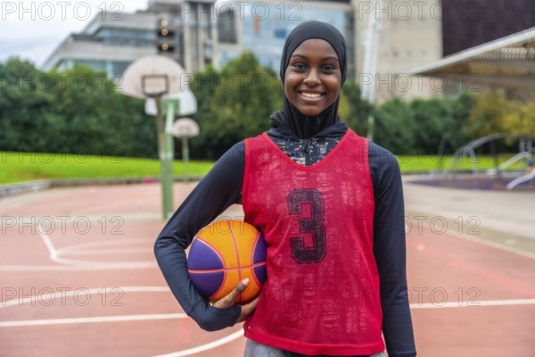 Young muslim woman wearing a hijab and sports uniform, holding a basketball and smiling confidently on an outdoor court, representing diversity in sport and active lifestyle