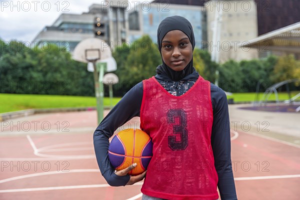 Young muslim woman wearing a hijab, long sleeve shirt, and basketball bib, holding a basketball while standing on an outdoor court, embodying confidence and athletic spirit