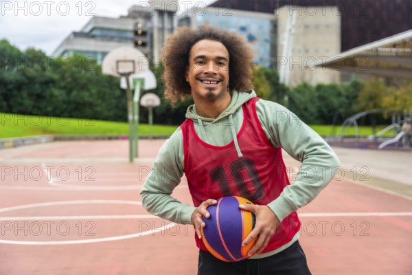Young man with afro hairstyle smiling and holding a colorful basketball while standing on an outdoor urban basketball court, showing a positive and healthy lifestyle