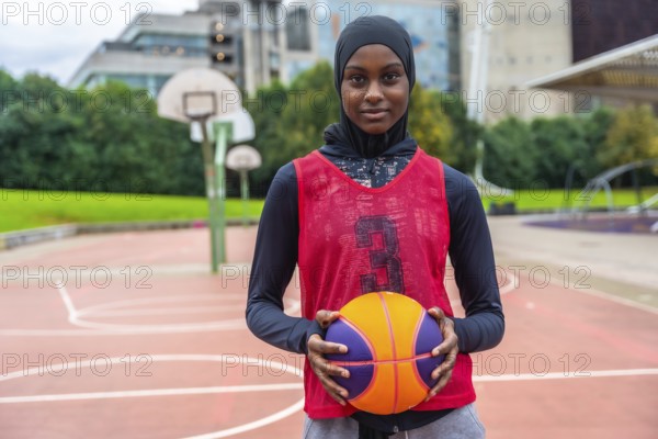 Young woman in a hijab and sports vest standing on an outdoor basketball court, holding a colorful basketball while looking directly at the camera, symbolizing empowerment and athletic diversity