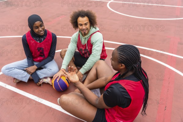 Diverse young friends taking a break on an outdoor basketball court, sitting and talking while holding a ball, enjoying their shared pastime and company