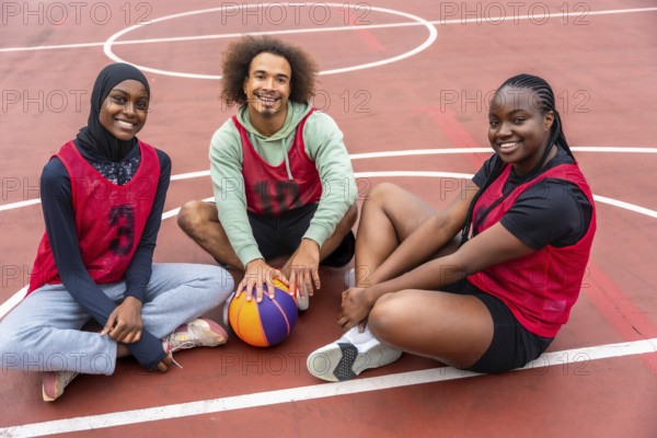 Diverse young friends wearing sport jerseys and a hijab, smiling while sitting together on an outdoor basketball court with a ball, representing teamwork and active youth