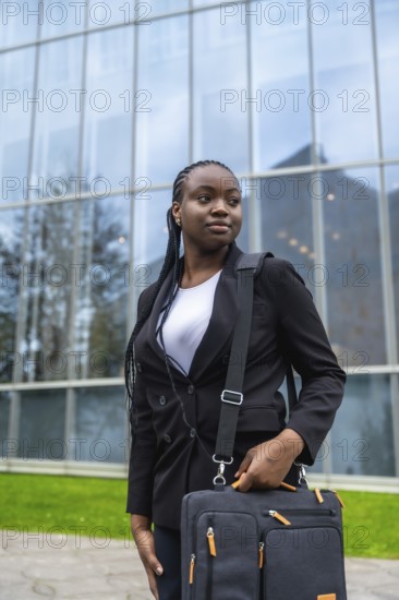 Confident black businesswoman standing outdoors, holding a briefcase, looking away while waiting, symbolizing professionalism, success, and ambition in a corporate urban environment