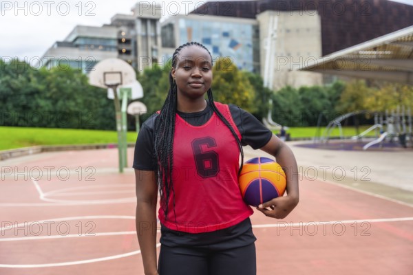 Young black woman wearing a red number six jersey and black athletic attire, standing confidently on an outdoor basketball court while holding a colorful basketball