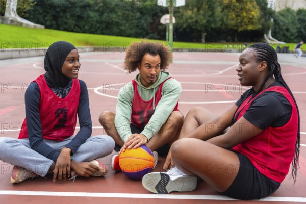 Diverse young adult friends wearing athletic jerseys sitting on an urban outdoor basketball court, happily engaging in conversation and fostering positive relationships within their community