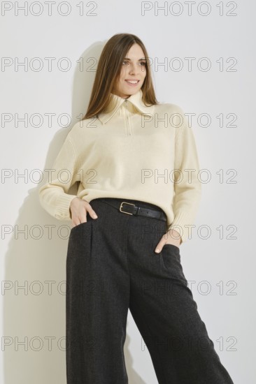 A woman leaning on bright wall in a trendy outfit consisting of a light sweater and dark wide-leg pants