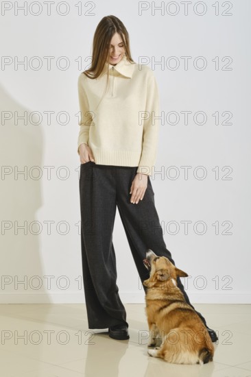 Stylish woman wearing a light sweater and wide-leg pants stands indoors, playfully engaging with a friendly dog