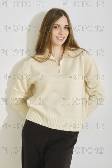 A young woman poses in a stylish, cream-colored sweater paired with black pants. She looks with playful smile while holding both hands behind her back
