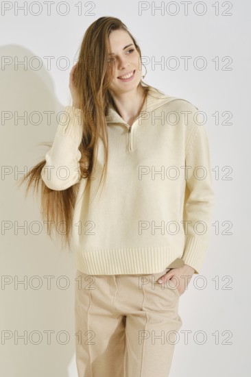 A woman stands in a modern indoor space, smiling with her long hair flowing. She wears a light, cozy sweater paired with relaxed tan pants, embodying a balance of fashion and practicality