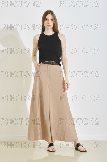 A young woman showcases a stylish outfit with a fitted black tank top and elegant beige wide-leg pants. She stands confidently in a bright, minimalist studio, perfect for practical fashion