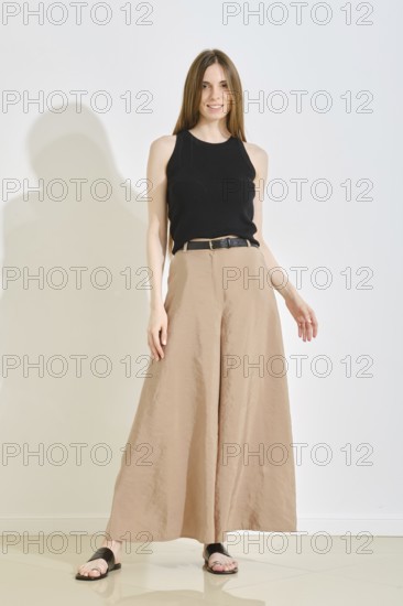 A young woman stands confidently in a trendy outfit, showcasing wide-legged beige pants and a black sleeveless top. This stylish look emphasizes comfort and practicality, perfect for casual settings