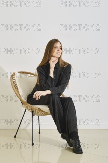 A young woman wears a oversized black outfit while seated comfortably in a modern chair. Her relaxed pose highlights the practicality of her attire in a bright, minimalistic indoor space